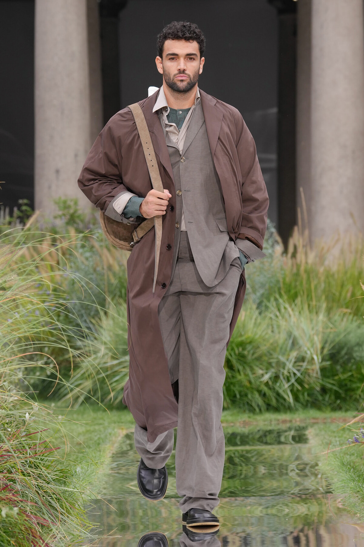 Male model walking outdoors in a brown trench coat over a gray suit, carrying a tan shoulder bag.