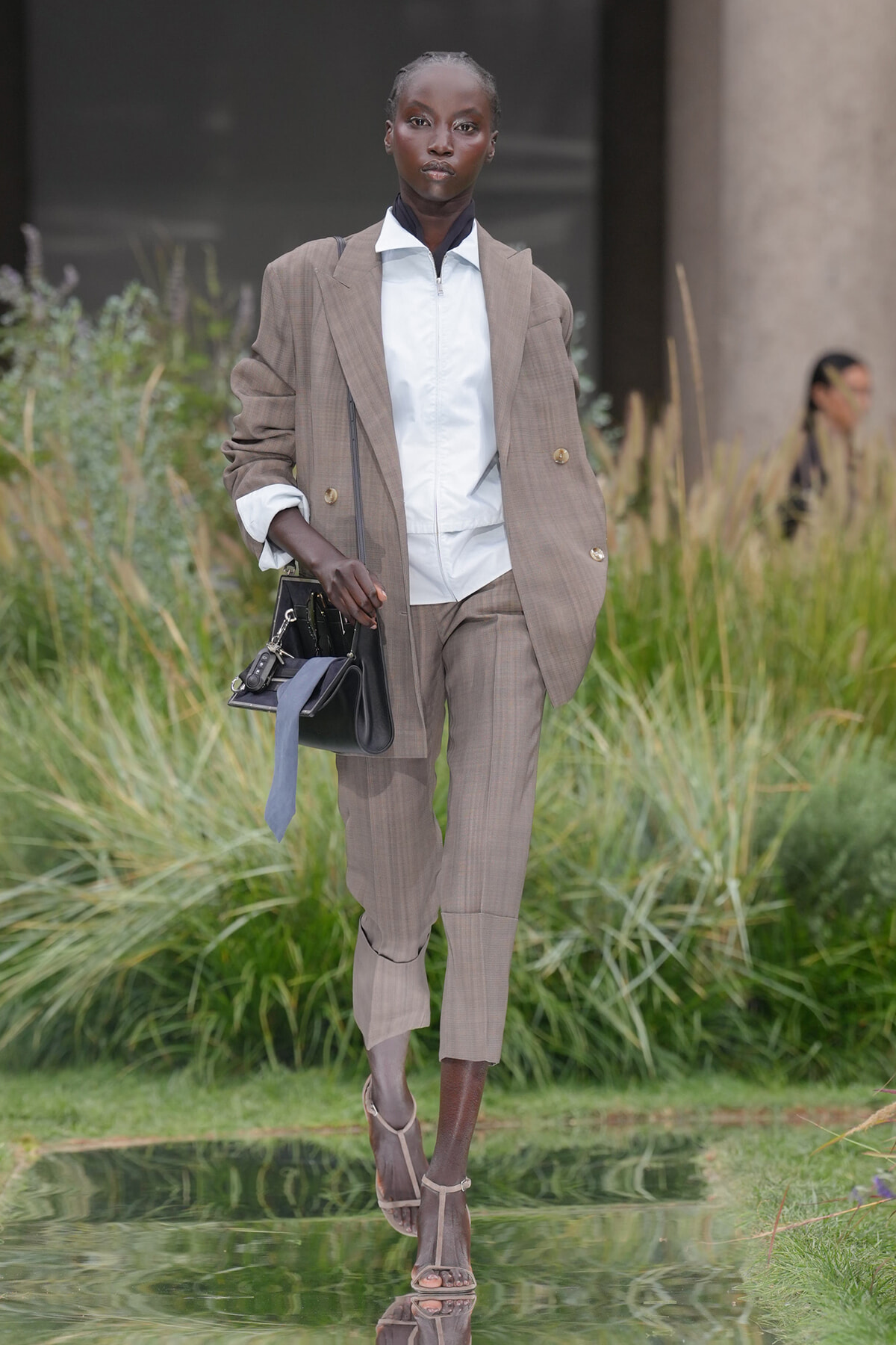 Model walking a runway in a taupe suit: oversized blazer, white zip-front shirt, matching cropped trousers, carrying a black handbag with a light blue scarf tied to it, tan heeled sandals, outdoors in a grassy setting