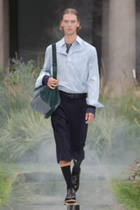 Male model walking on a runway in a light blue shirt, dark navy cropped pants, black belt, black shoes, and a large green-gray shoulder bag.