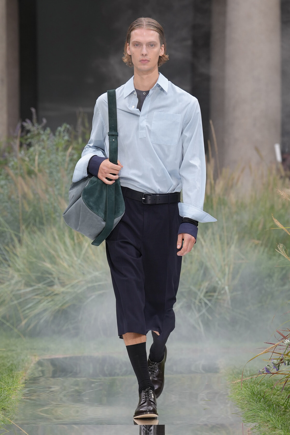 Male model walking on a runway in a light blue shirt, dark navy cropped pants, black belt, black shoes, and a large green-gray shoulder bag.