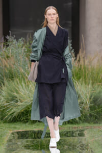 Model walking outdoors in a navy wrap suit with a green trench draped over shoulders, holding a beige bag, wearing white mules.