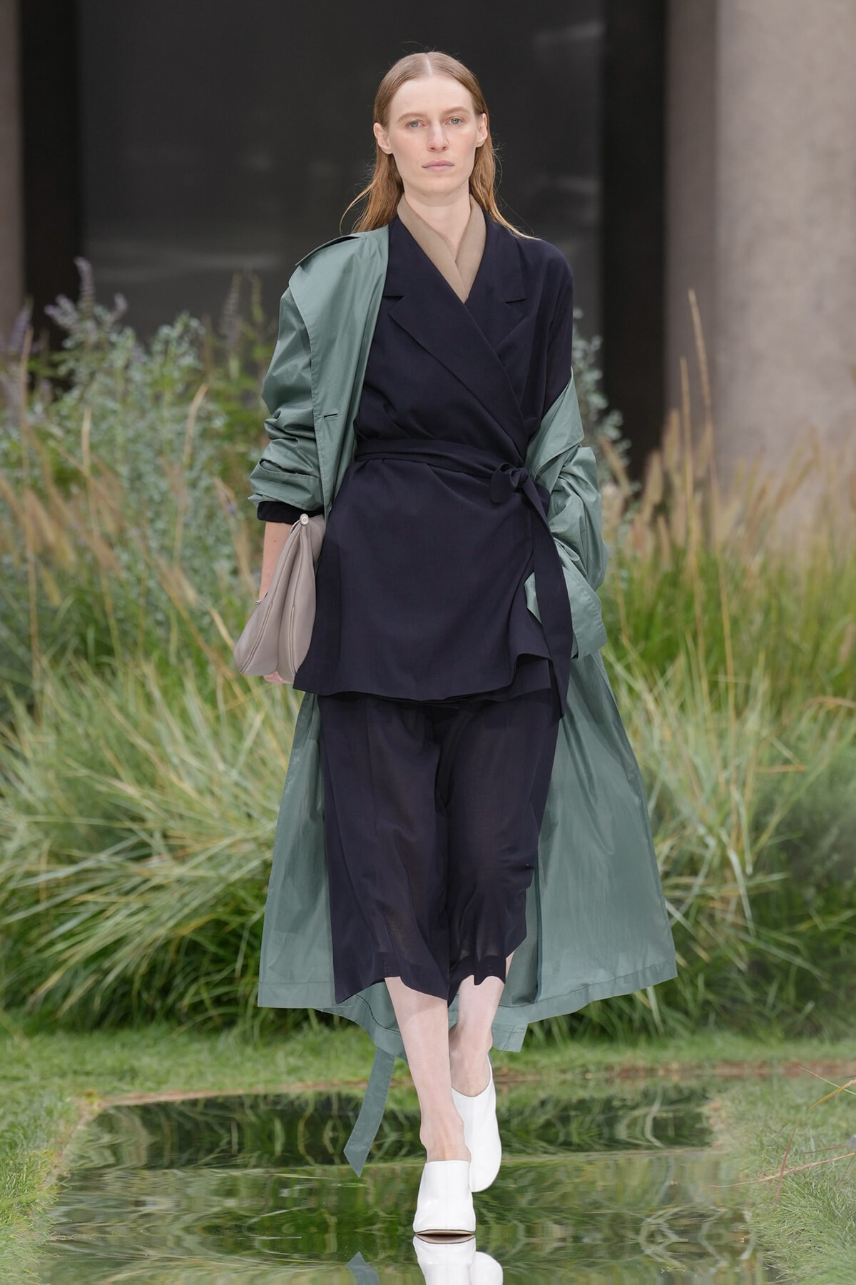 Model walking outdoors in a navy wrap suit with a green trench draped over shoulders, holding a beige bag, wearing white mules.