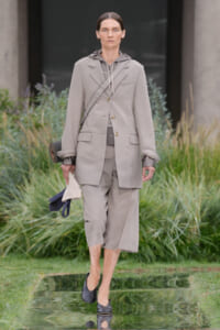 Model walking outdoors in a light gray suit: oversized blazer with a crossbody strap, matching cropped pants, hoodie underneath, and rolled cuffs with a rolled clutch and bag.