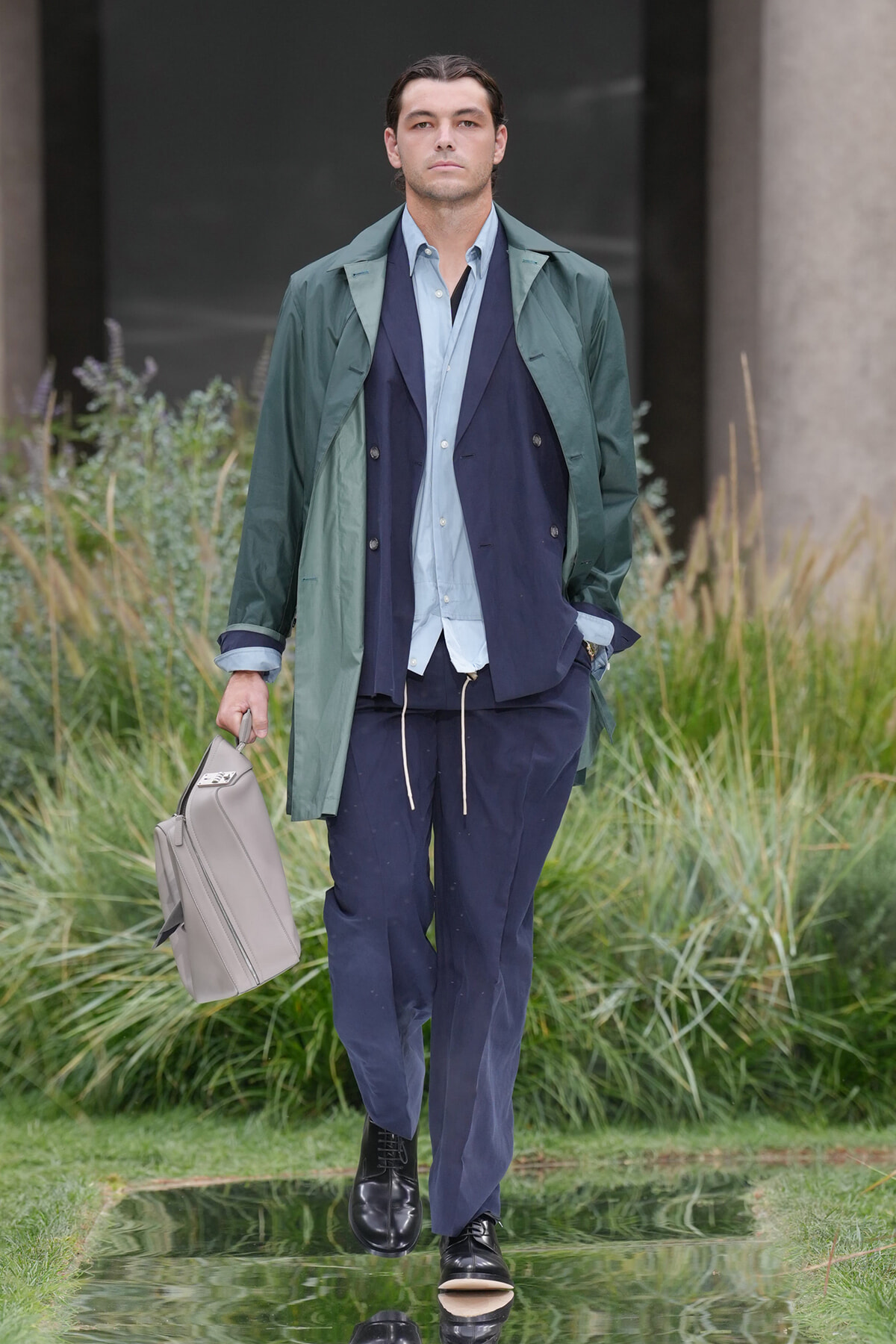 Man walking outdoors in layered blue suit pieces, green trench coat, and carrying a gray briefcase.