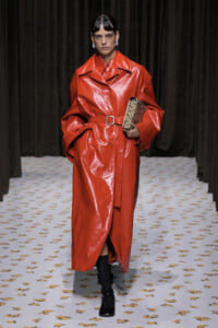 Model walking a runway in a red shiny trench coat with a belt, holding a snakeskin clutch and black ankle boots.