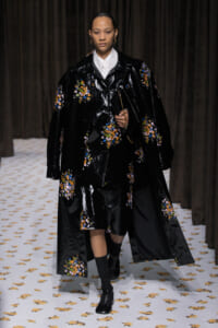 Runway model wearing a glossy black floral-embroidered coat over a white shirt, holding a black clutch, walking on a fashion show.