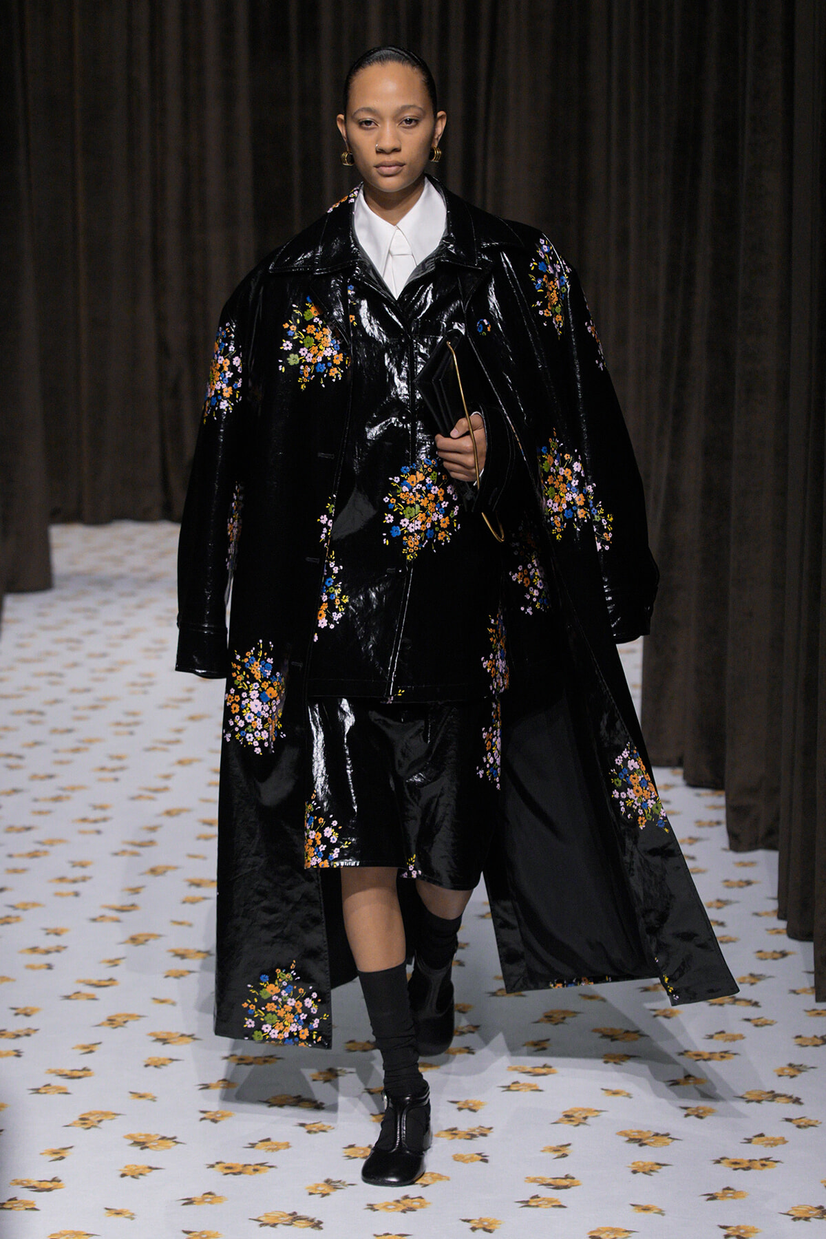 Runway model wearing a glossy black floral-embroidered coat over a white shirt, holding a black clutch, walking on a fashion show.