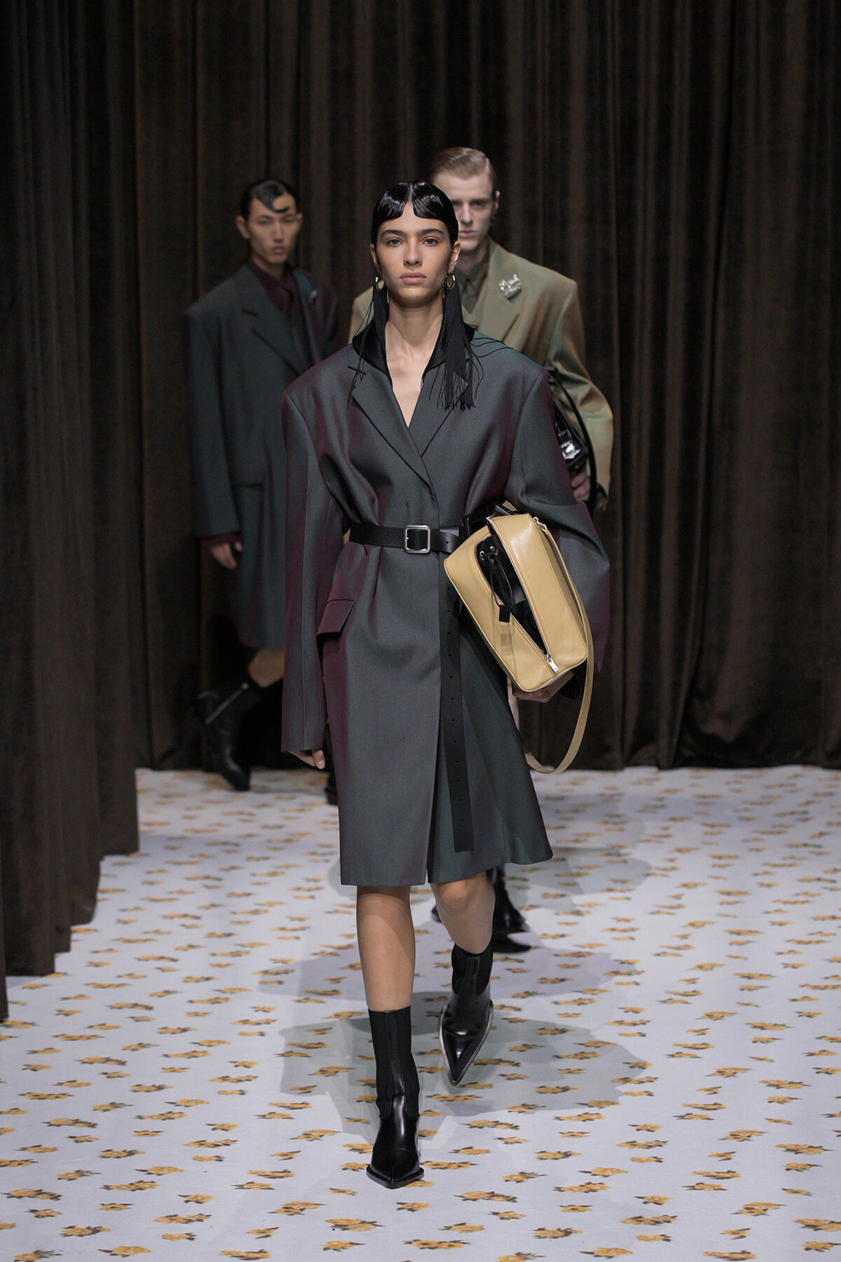 Model walks a fashion runway wearing a dark gray belted coat with a tan handbag and black boots; black fringe earrings, curtain backdrop.