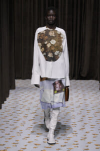Model wearing a white long-sleeve top with a floral patterned bib, paired with a printed skirt and white knee-high boots on a patterned runway.