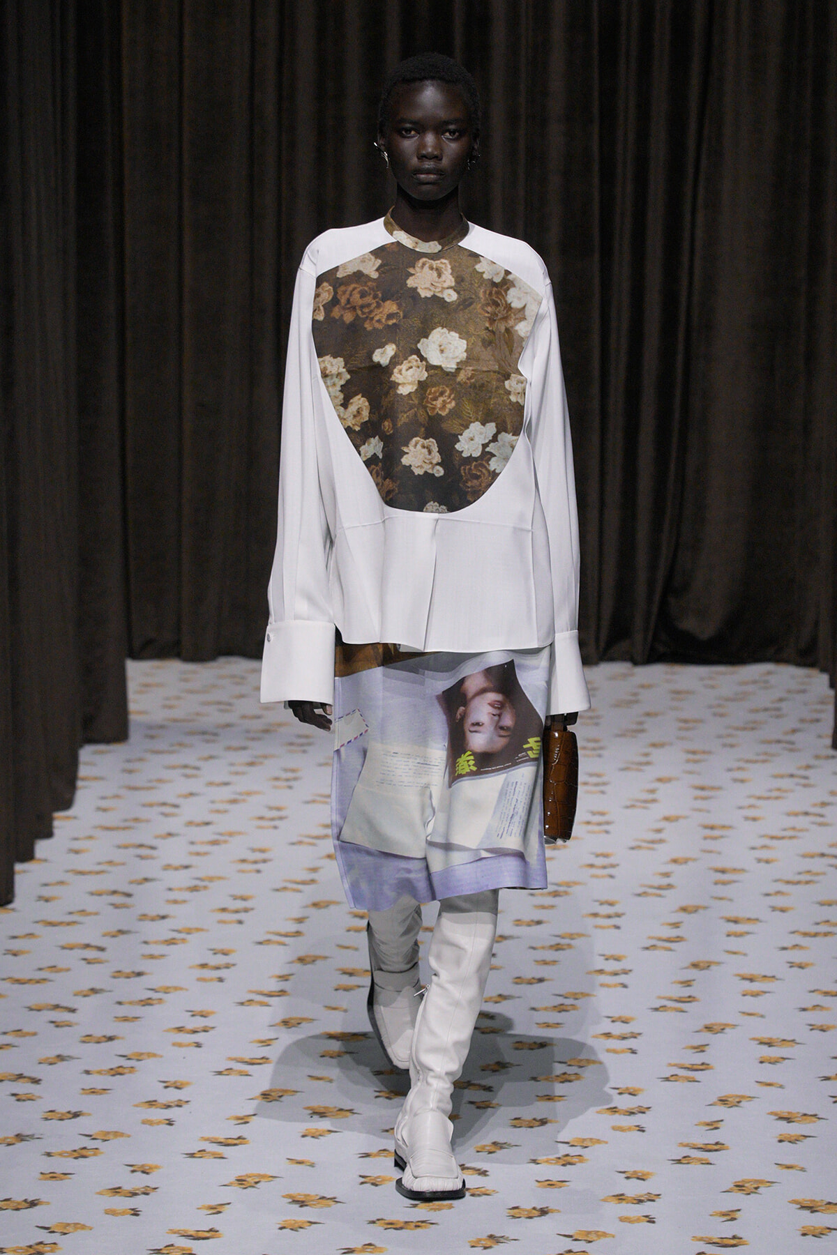 Model wearing a white long-sleeve top with a floral patterned bib, paired with a printed skirt and white knee-high boots on a patterned runway.