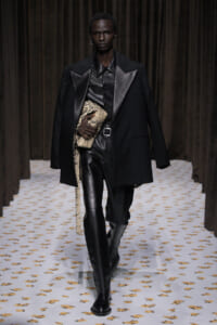 Male model walks the runway in a black leather-on-leather outfit with an oversized black coat, clutching a snakeskin bag