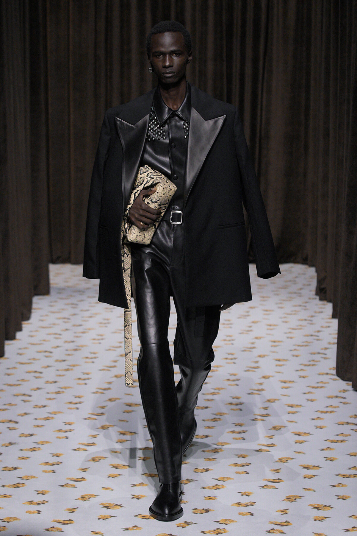 Male model walks the runway in a black leather-on-leather outfit with an oversized black coat, clutching a snakeskin bag