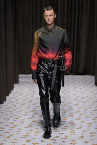 Male model walking runway in a gradient green-to-red shirt with black patent leather pants and boots, holding a black clutch/strap bag at the side