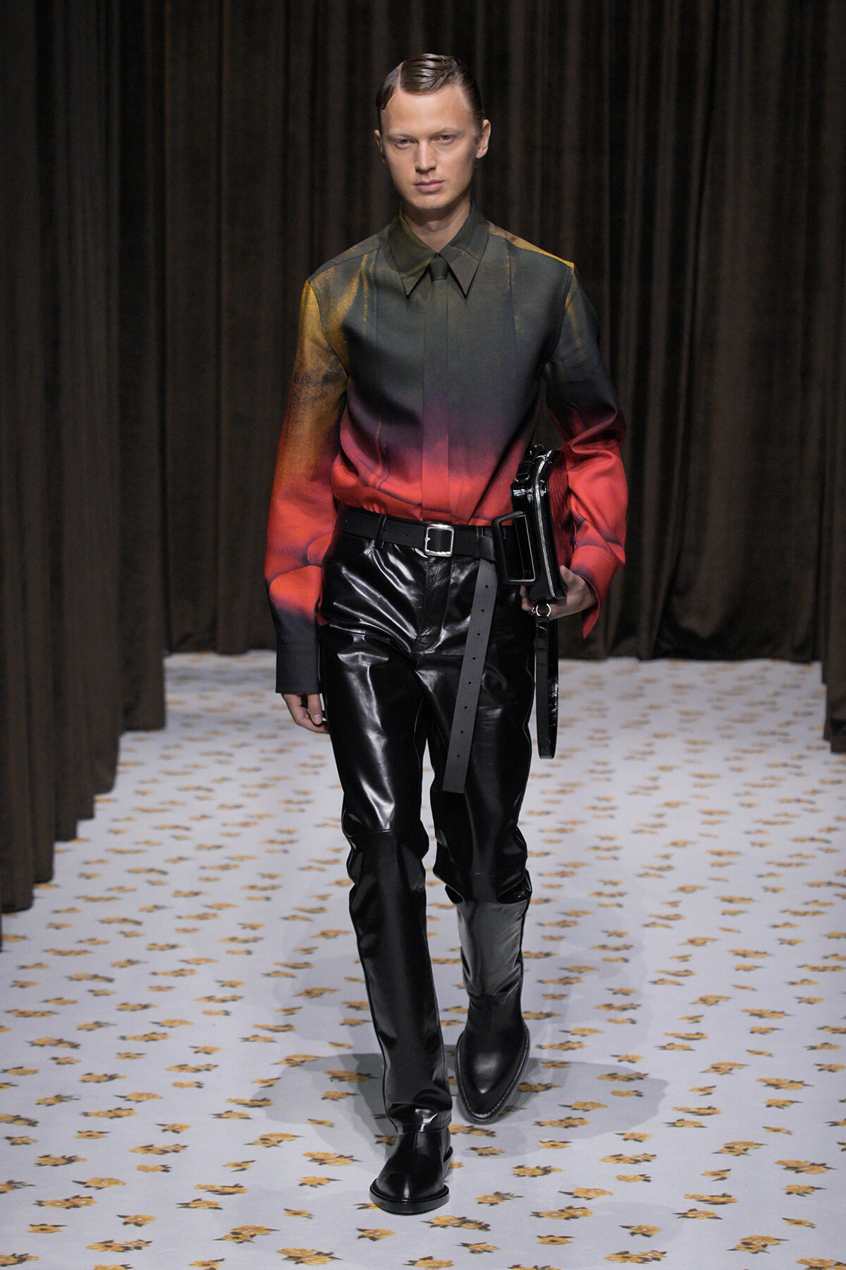 Male model walking runway in a gradient green-to-red shirt with black patent leather pants and boots, holding a black clutch/strap bag at the side