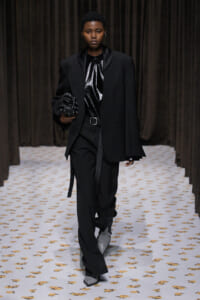 Model on a runway in an oversized black blazer, shiny black top, black trousers with a belt, gray boots, and a studded black handbag.