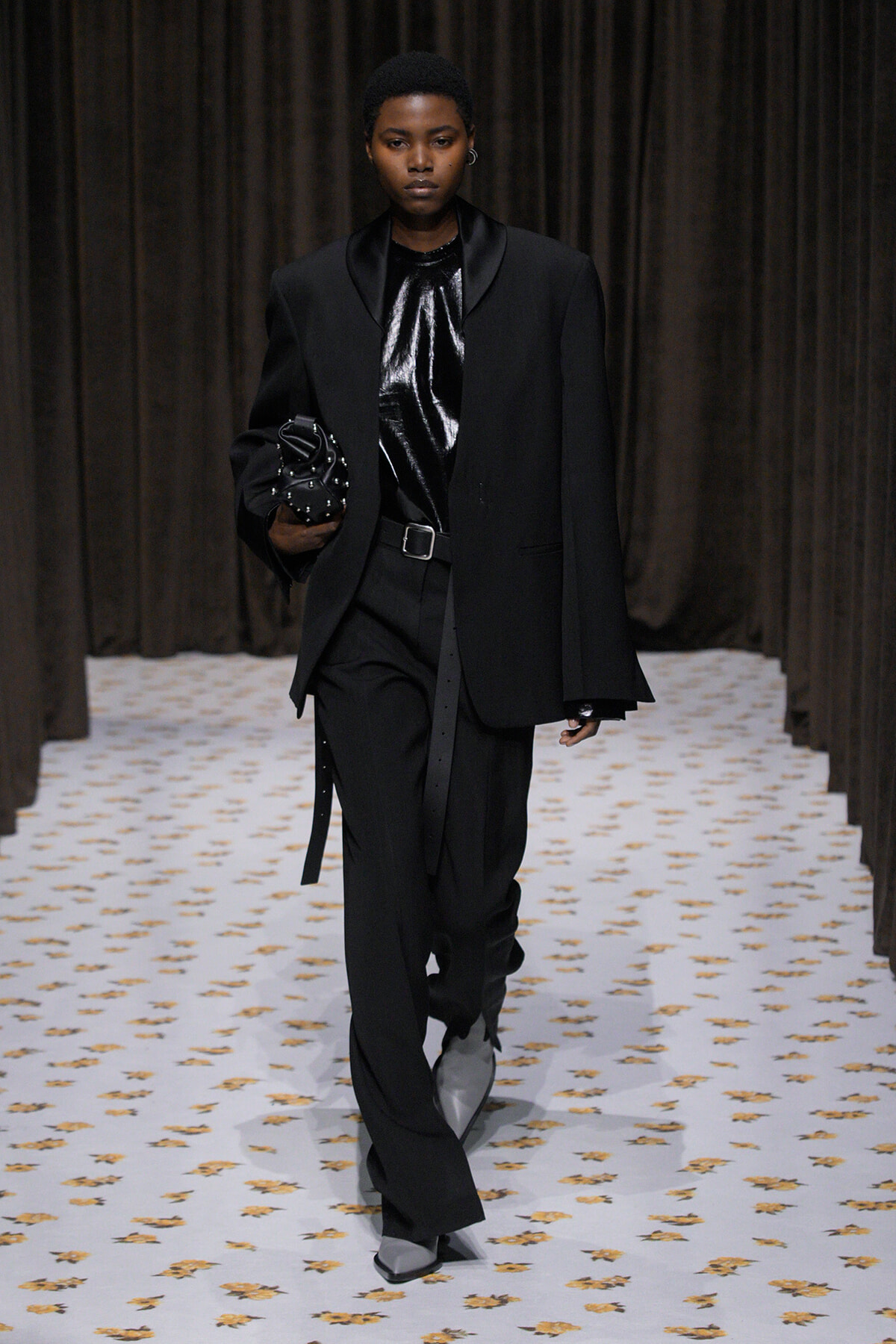 Model on a runway in an oversized black blazer, shiny black top, black trousers with a belt, gray boots, and a studded black handbag.