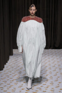 Model walks a runway in a voluminous white dress with a red sequined yoke and white ankle boots.