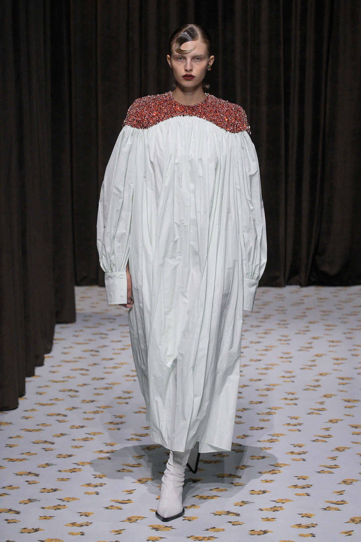 Model walks a runway in a voluminous white dress with a red sequined yoke and white ankle boots.