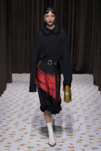 Model on a runway wearing a black blouse with a ruffled collar, belted waist, and a gradient red-to-black midi skirt, with white boots and a mustard clutch.