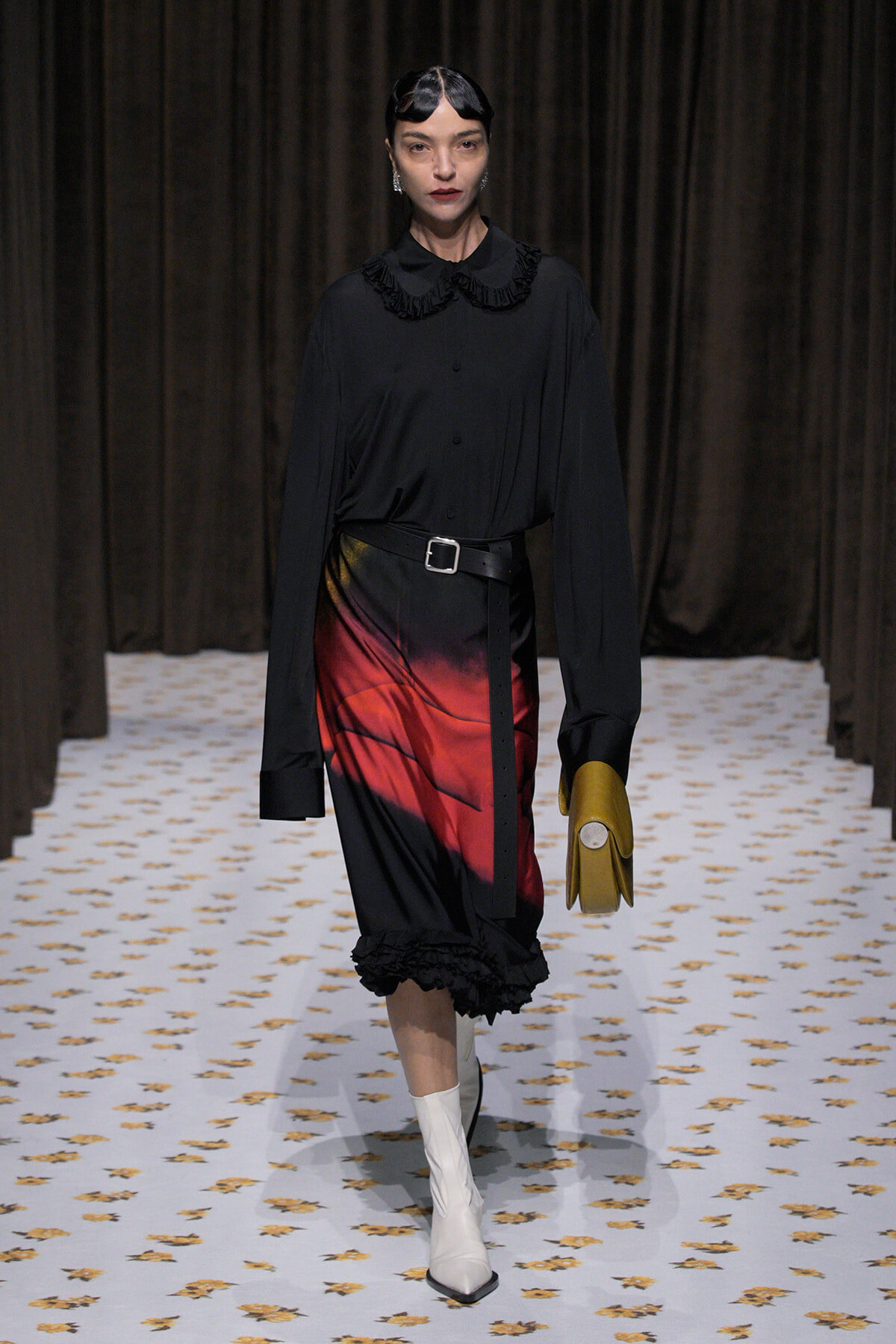 Model on a runway wearing a black blouse with a ruffled collar, belted waist, and a gradient red-to-black midi skirt, with white boots and a mustard clutch.
