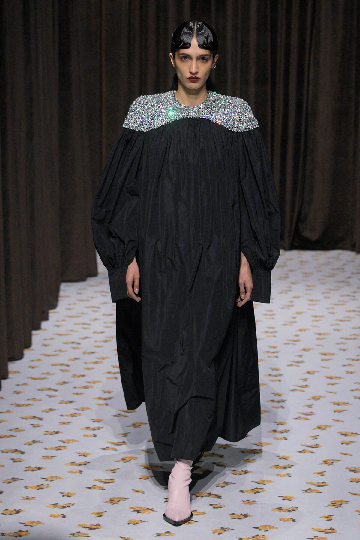 Model in a sweeping black gown with a sparkling silver embellished yoke and pale pink ankle boots on the runway.