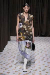 Model walking runway in a sleeveless floral-print dress with a collage panel at the hem and white knee-high boots, holding a small black bag.