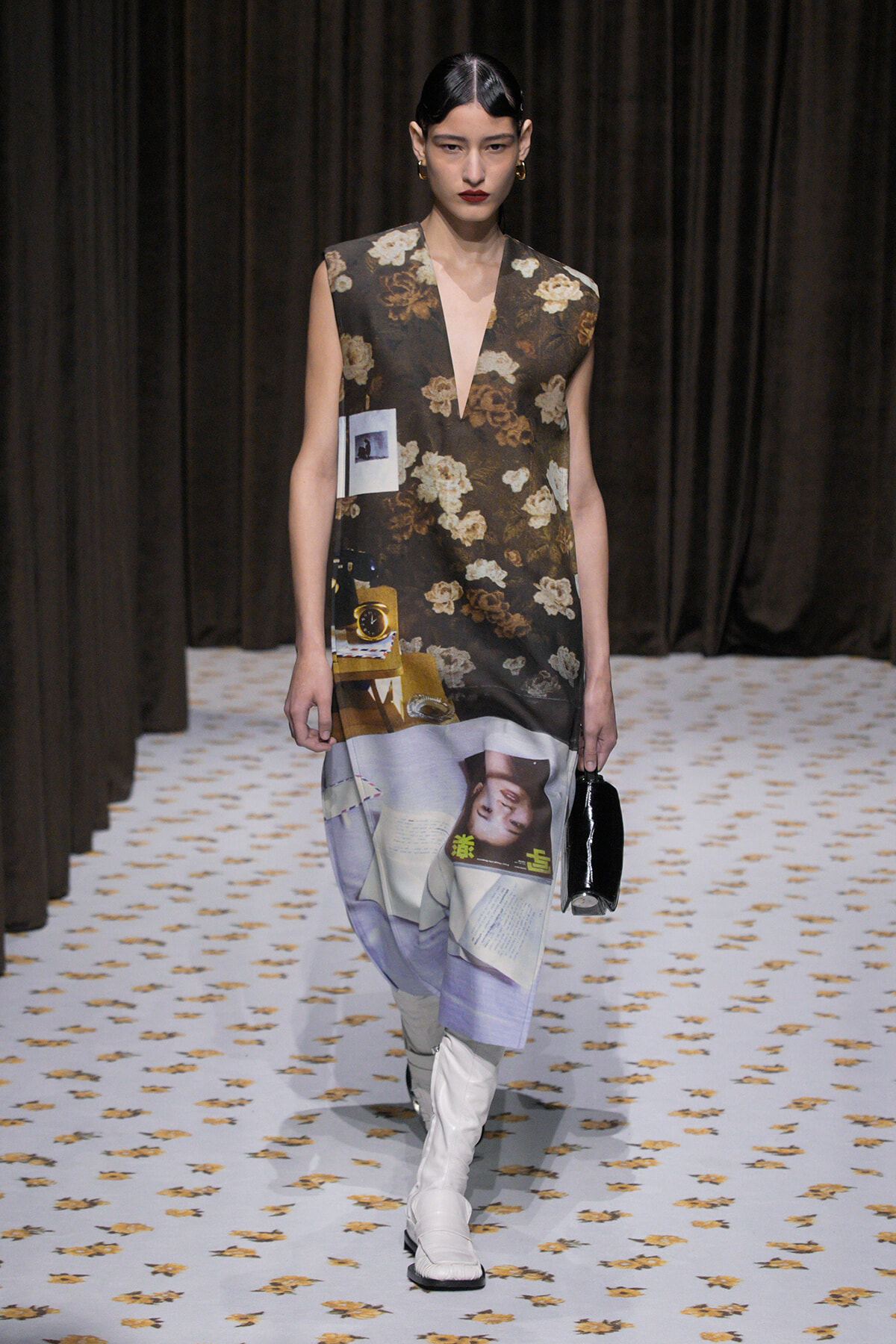 Model walking runway in a sleeveless floral-print dress with a collage panel at the hem and white knee-high boots, holding a small black bag.