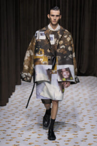 Male model walks the runway in a brown floral collage coat with photo-montage prints, white shirt, and black loafers.