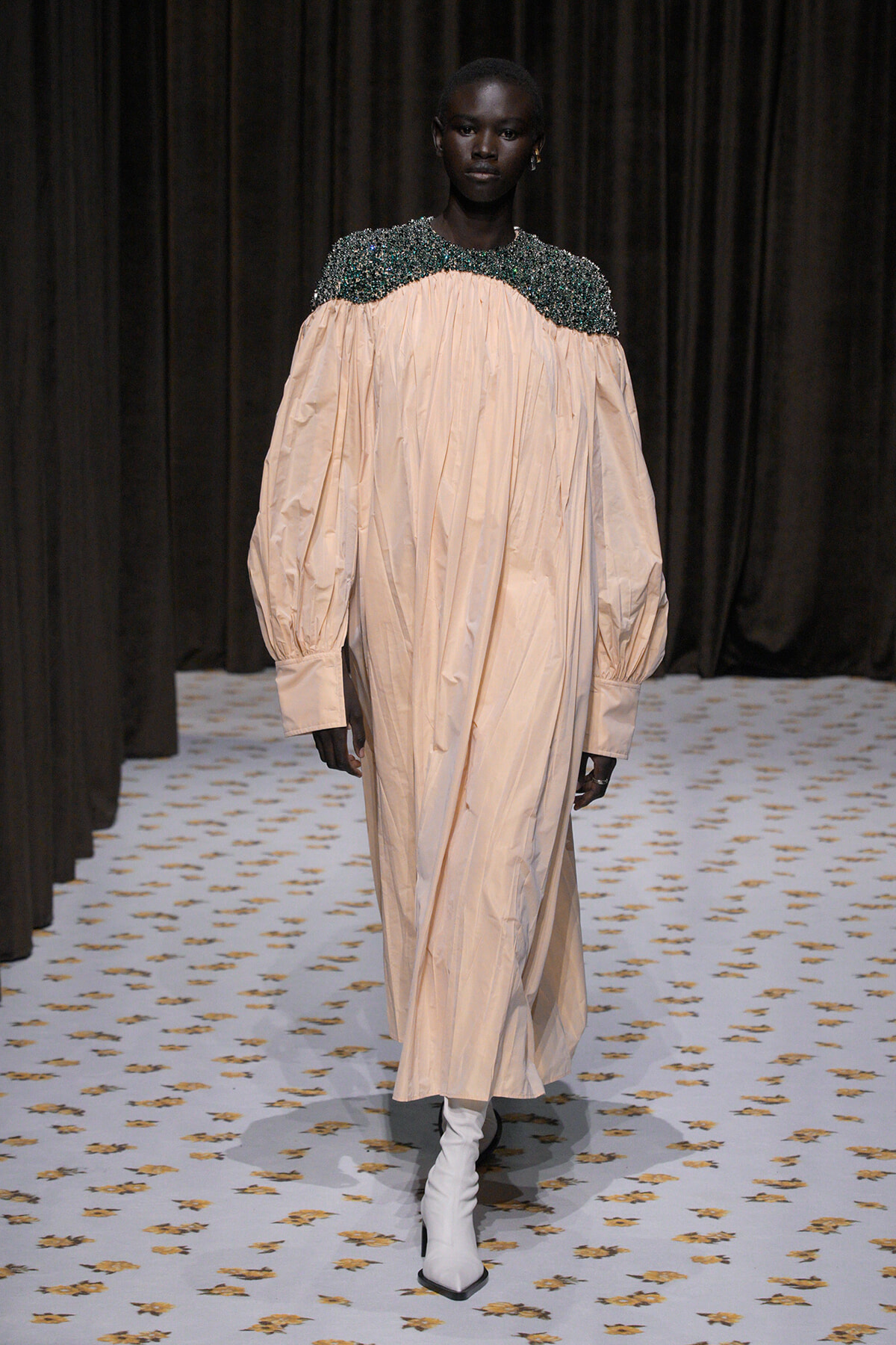 Model walking runway in a peach oversized gown with a beaded dark emerald collar and white ankle boots on a patterned floor.