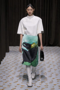 Model walking the runway in a white oversized top, mint-to-dark graphic skirt featuring a car print, white knee-high boots, and a small black handbag.