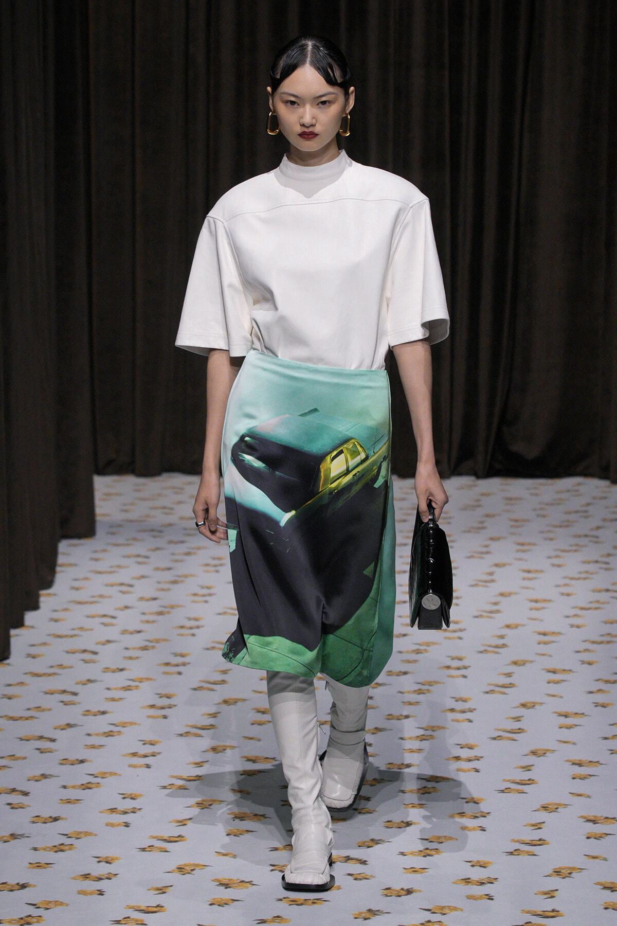 Model walking the runway in a white oversized top, mint-to-dark graphic skirt featuring a car print, white knee-high boots, and a small black handbag.