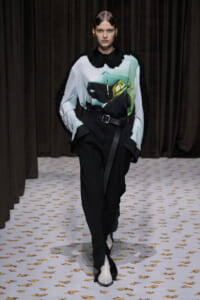 Model walking a fashion show wearing a light blue graphic blouse with a car image, black high-waisted pants with belt, and black fringed cape sleeves.