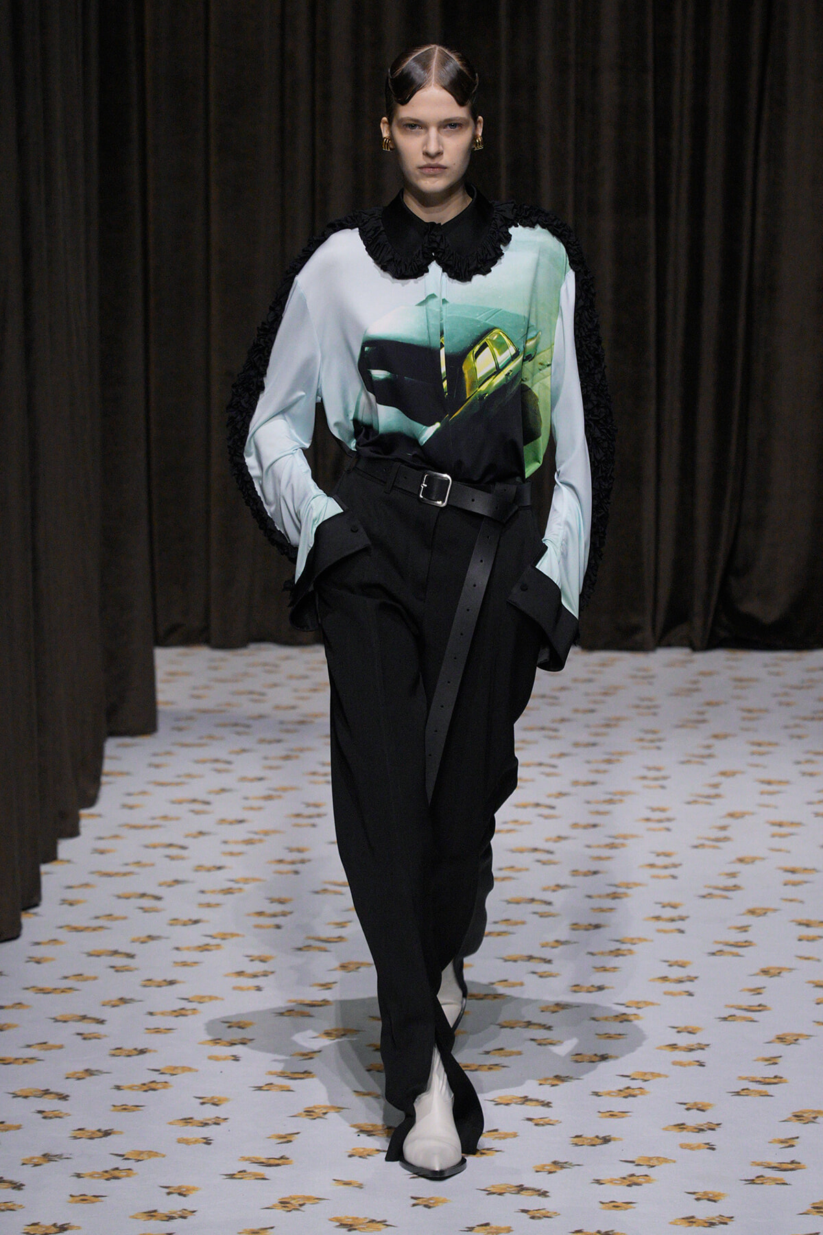 Model walking a fashion show wearing a light blue graphic blouse with a car image, black high-waisted pants with belt, and black fringed cape sleeves.