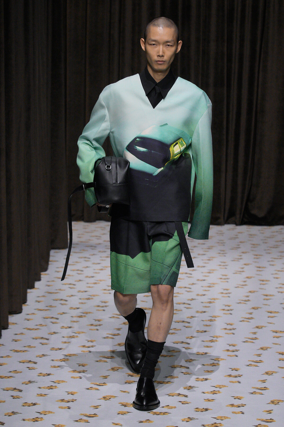 Runway model wearing a mint-green oversized top with a car graphic, green color-block shorts, black shoes, and a black shoulder bag.