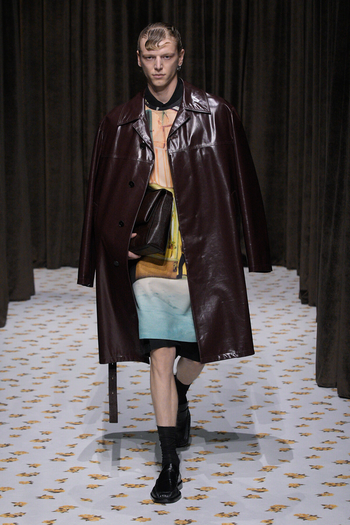 Runway model wearing an oversized burgundy leather trench over a colorful top and teal shorts, carrying a dark clutch.
