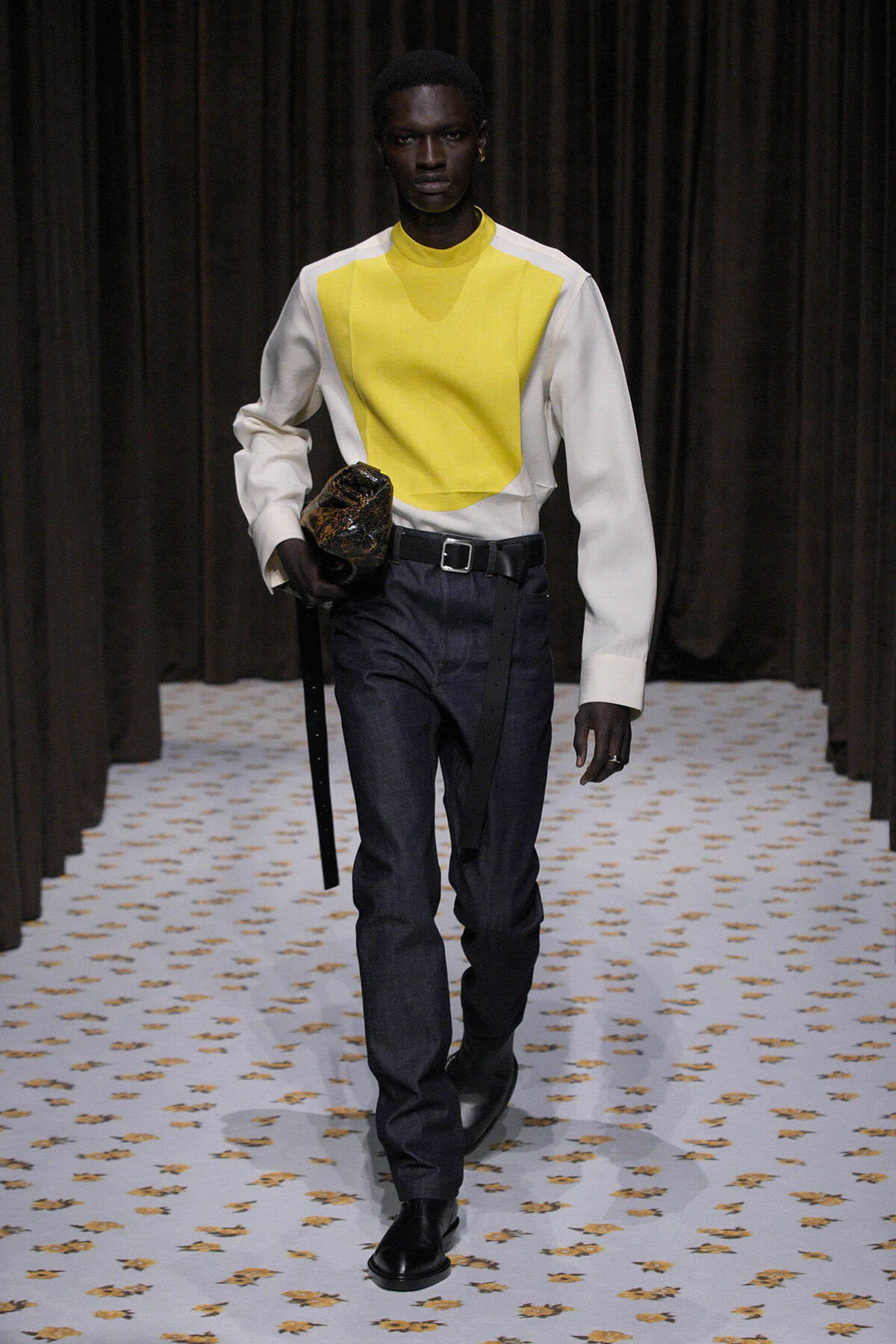 Male model walking a runway in a yellow and white color-block sweater, dark jeans, black belt, and boots, clutch in hand.