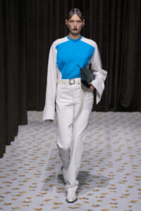 Model on a runway wearing a blue and white color-block top with white pants, a white belt, and a black clutch in hand.