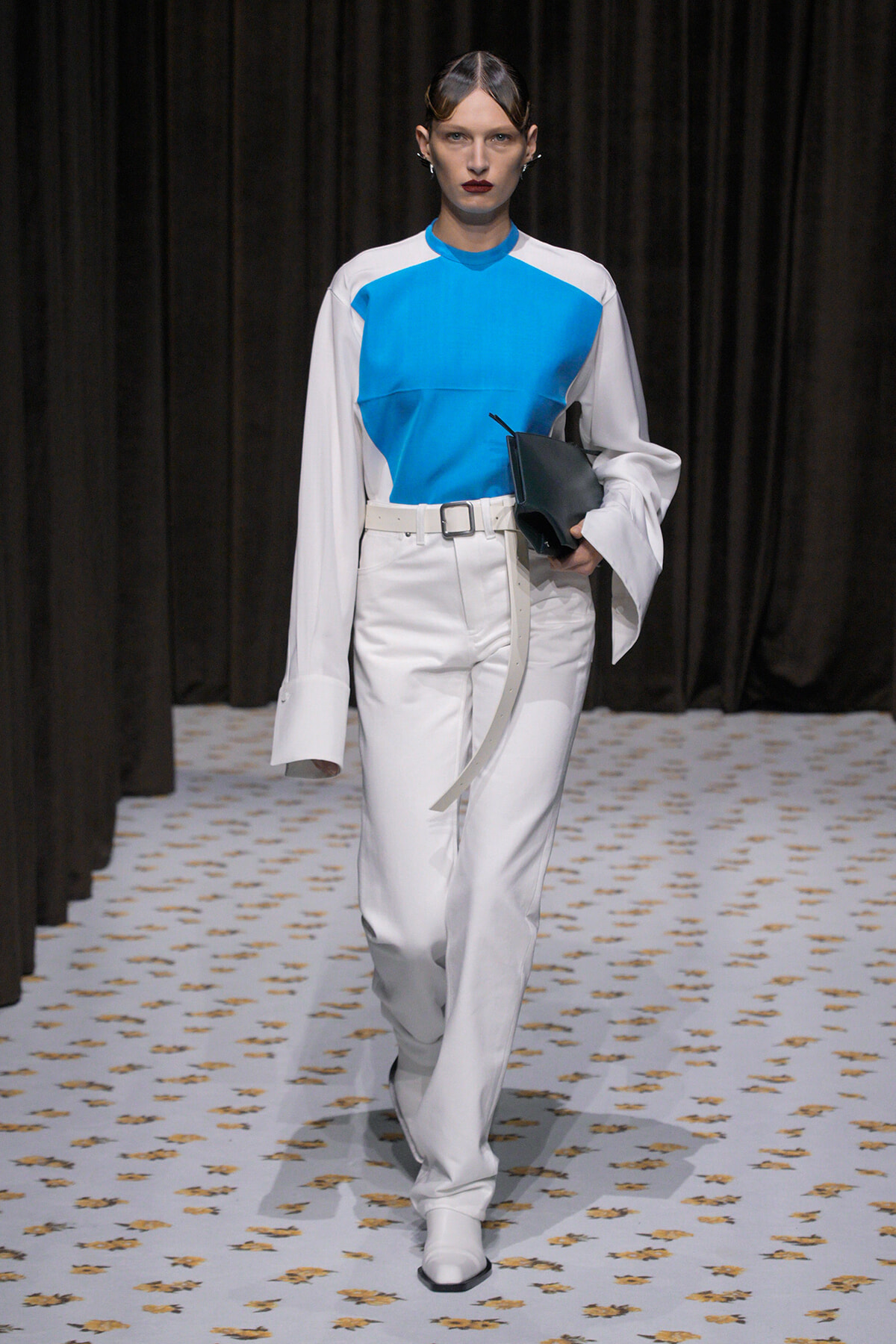 Model on a runway wearing a blue and white color-block top with white pants, a white belt, and a black clutch in hand.