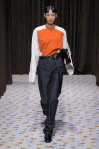 Model walks the runway in an orange-front color-block top with white sleeves, dark jeans, and a black belt while holding a black clutch.