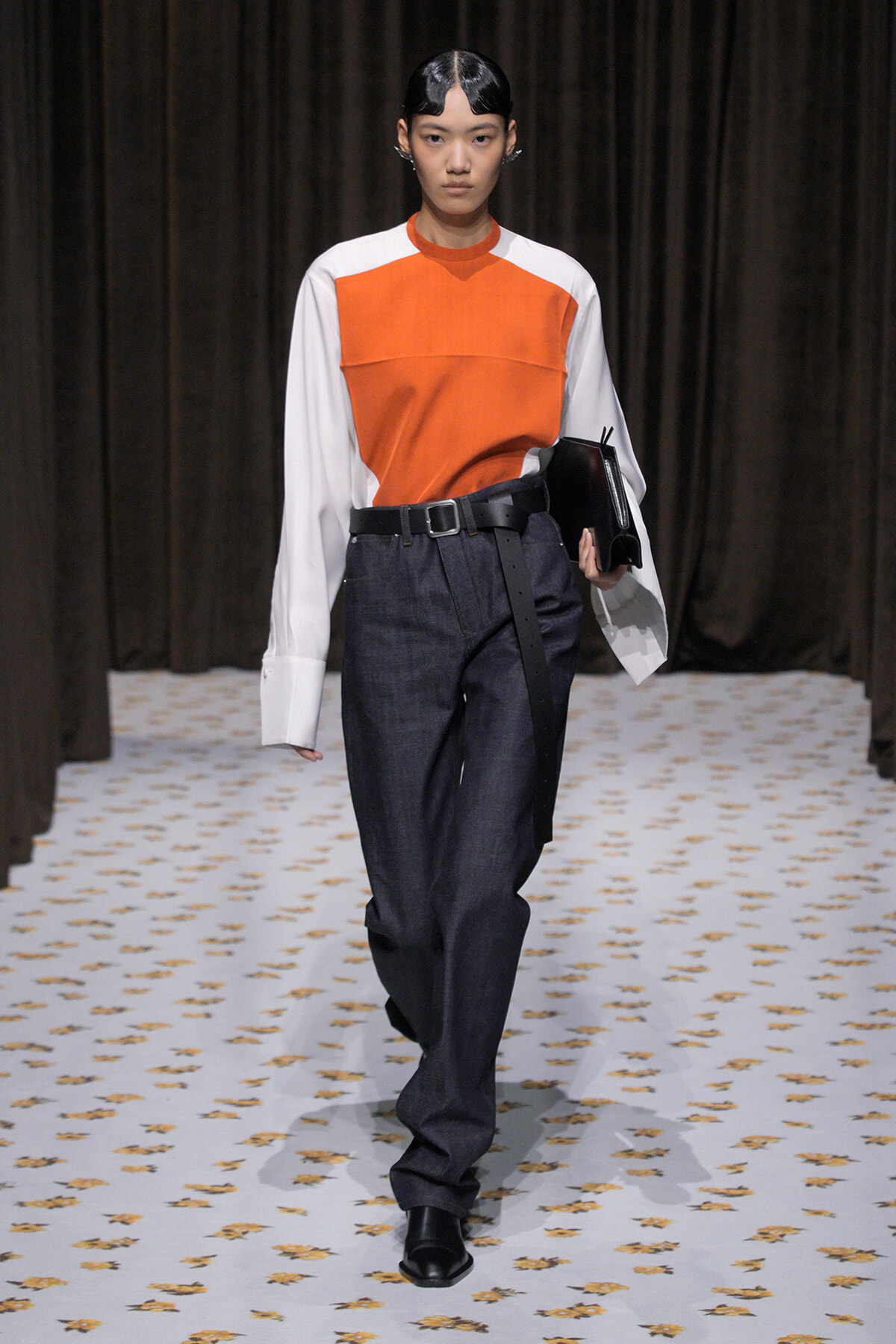 Model walks the runway in an orange-front color-block top with white sleeves, dark jeans, and a black belt while holding a black clutch.