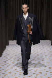 Male model walking runway in a dark blazer with satin lapels over a grey vest and white shirt, holding a brown rectangular bag.