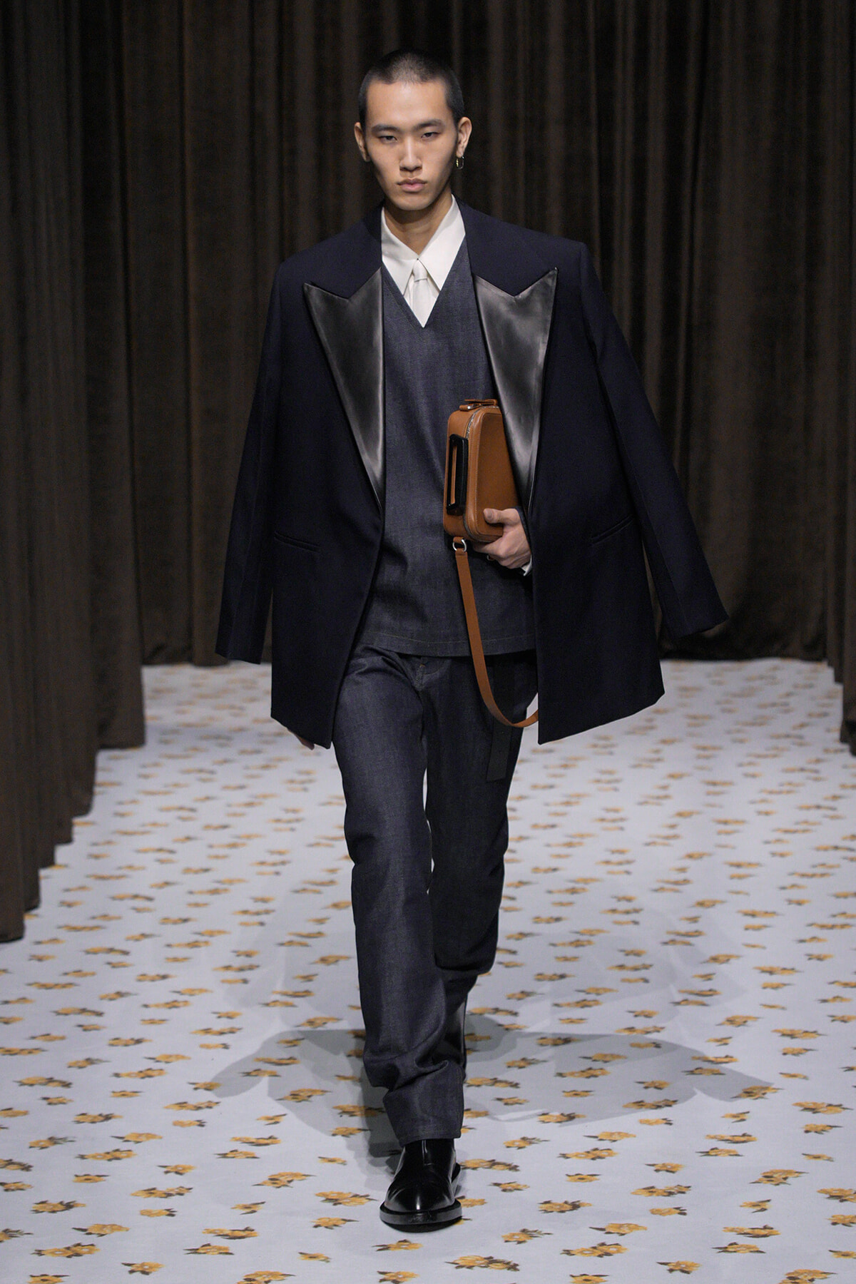 Male model walking runway in a dark blazer with satin lapels over a grey vest and white shirt, holding a brown rectangular bag.