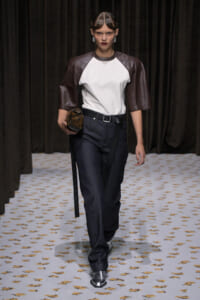 Runway model wearing a white front top with brown leather sleeves, dark jeans, a belt, and metallic boots, carrying a textured clutch.