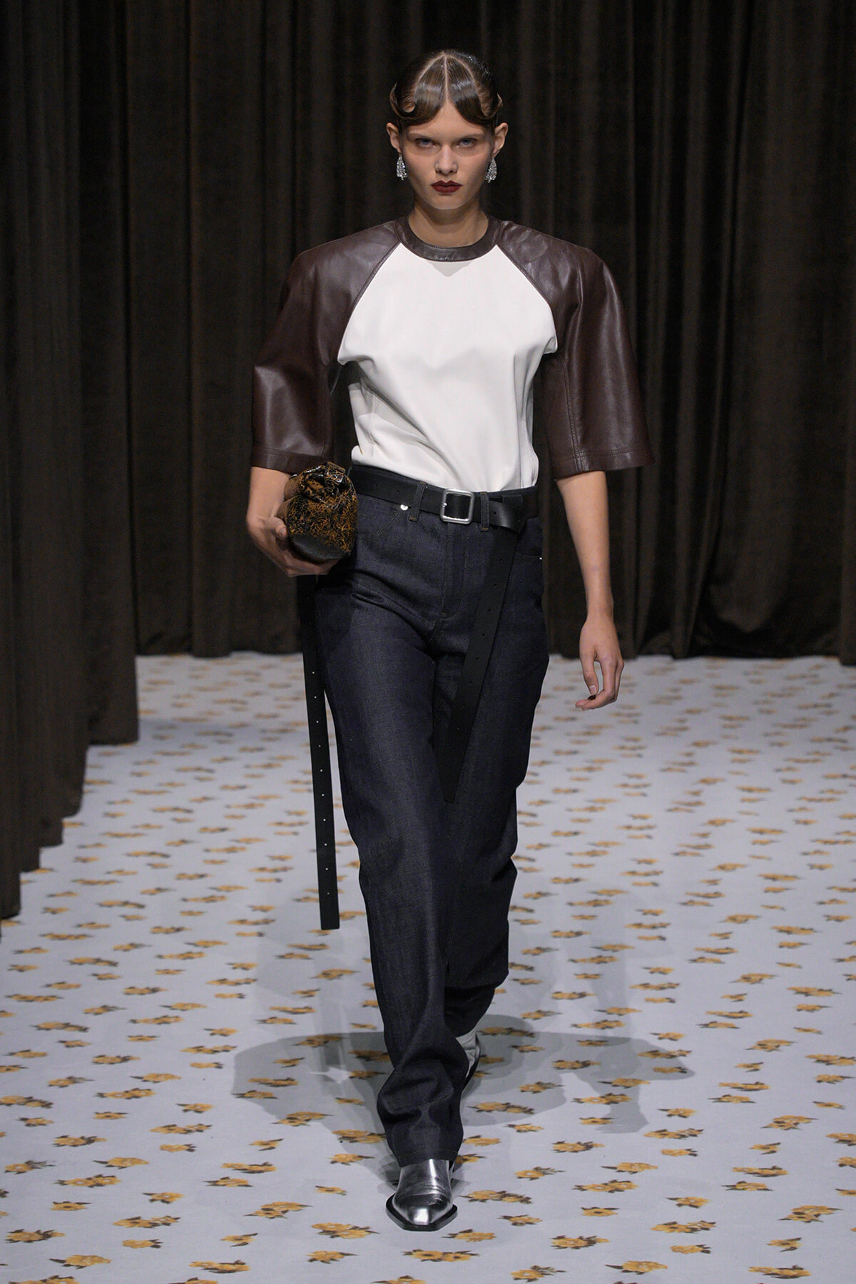 Runway model wearing a white front top with brown leather sleeves, dark jeans, a belt, and metallic boots, carrying a textured clutch.