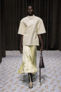 Model on a runway wearing an oversized cream top, pale satin skirt, snake-print boots, and carrying a brown bag.
