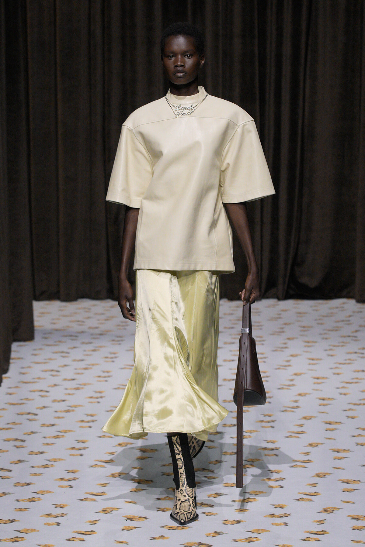 Model on a runway wearing an oversized cream top, pale satin skirt, snake-print boots, and carrying a brown bag.