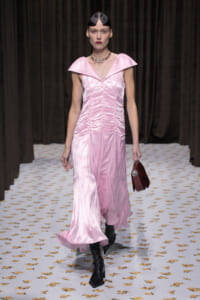 Model walking a fashion runway in a pink satin gown with a large flat collar and ruching, paired with black ankle boots and a burgundy handbag.