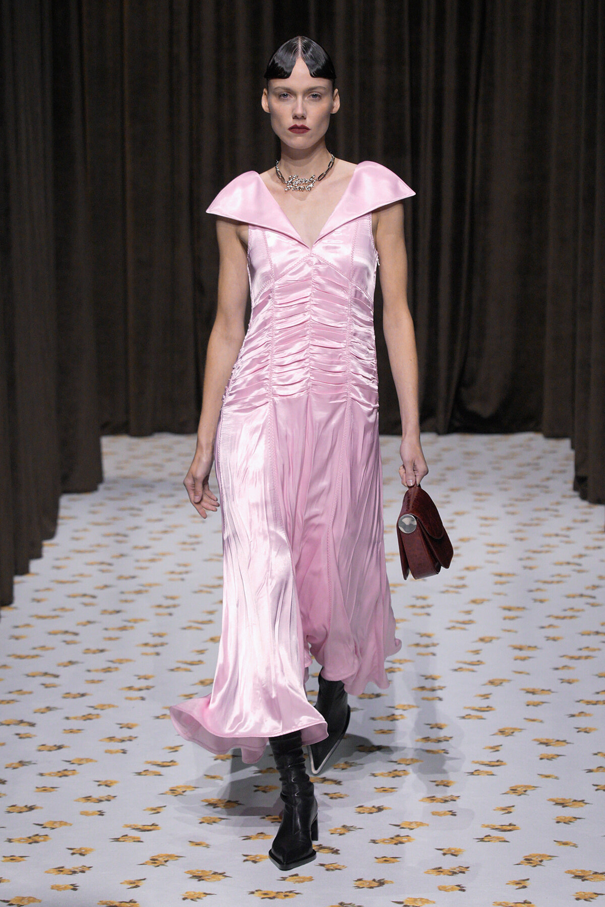 Model walking a fashion runway in a pink satin gown with a large flat collar and ruching, paired with black ankle boots and a burgundy handbag.