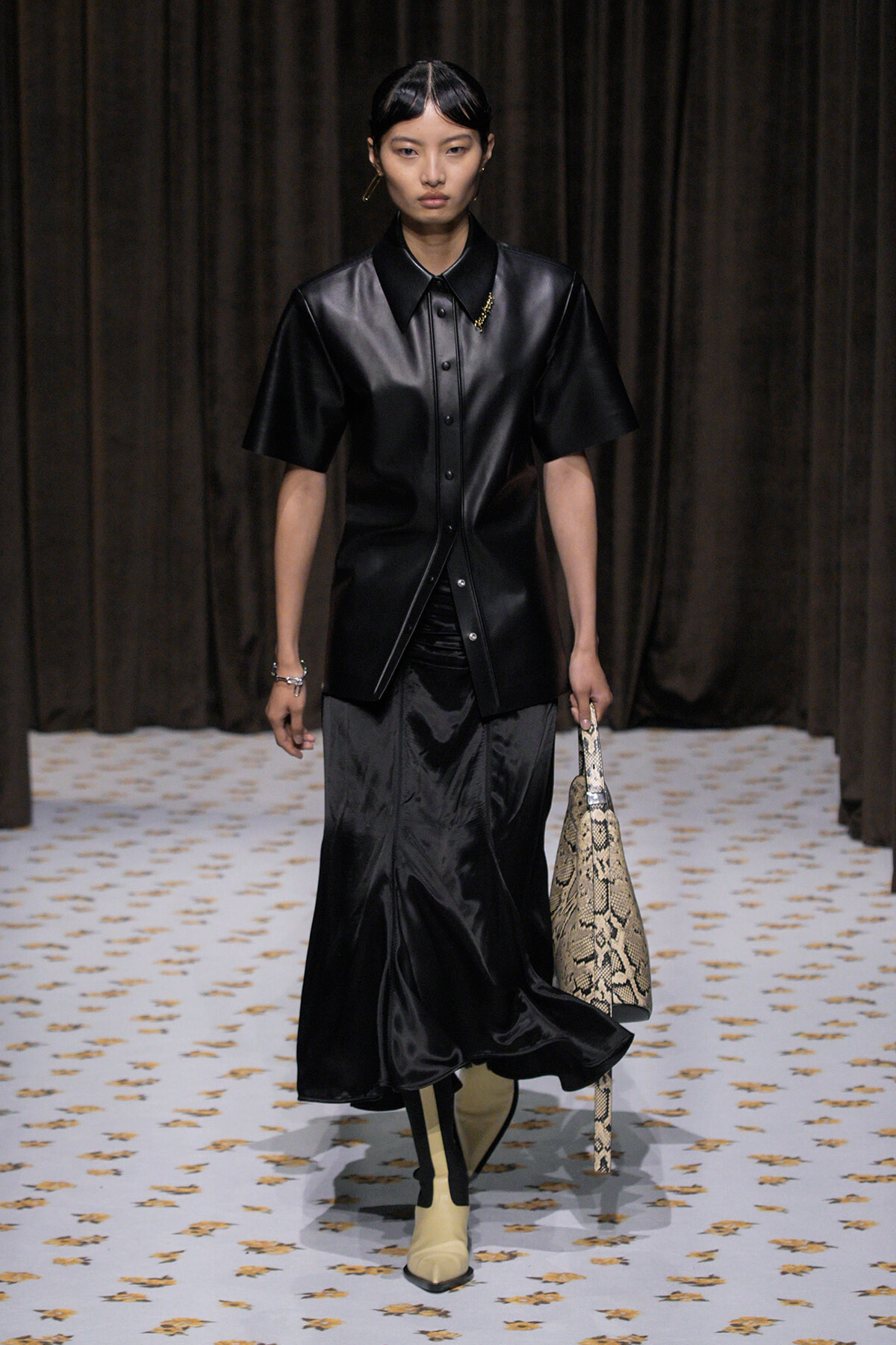 Model walking a runway in a black leather short-sleeve shirt, black satin midi skirt, beige boots, and a snakeskin handbag.