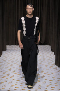 Male model on a fashion-show runway wearing a black top with white lace panels down the sides, black wide-leg trousers, and a black clutch/binder tucked under the arm.